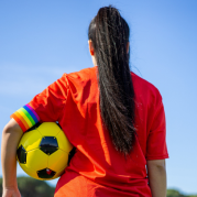 futebol feminino: mulher de costas, cabelos longos presos, vestindo camisa laranja e segurando uma bola de futebol amarela